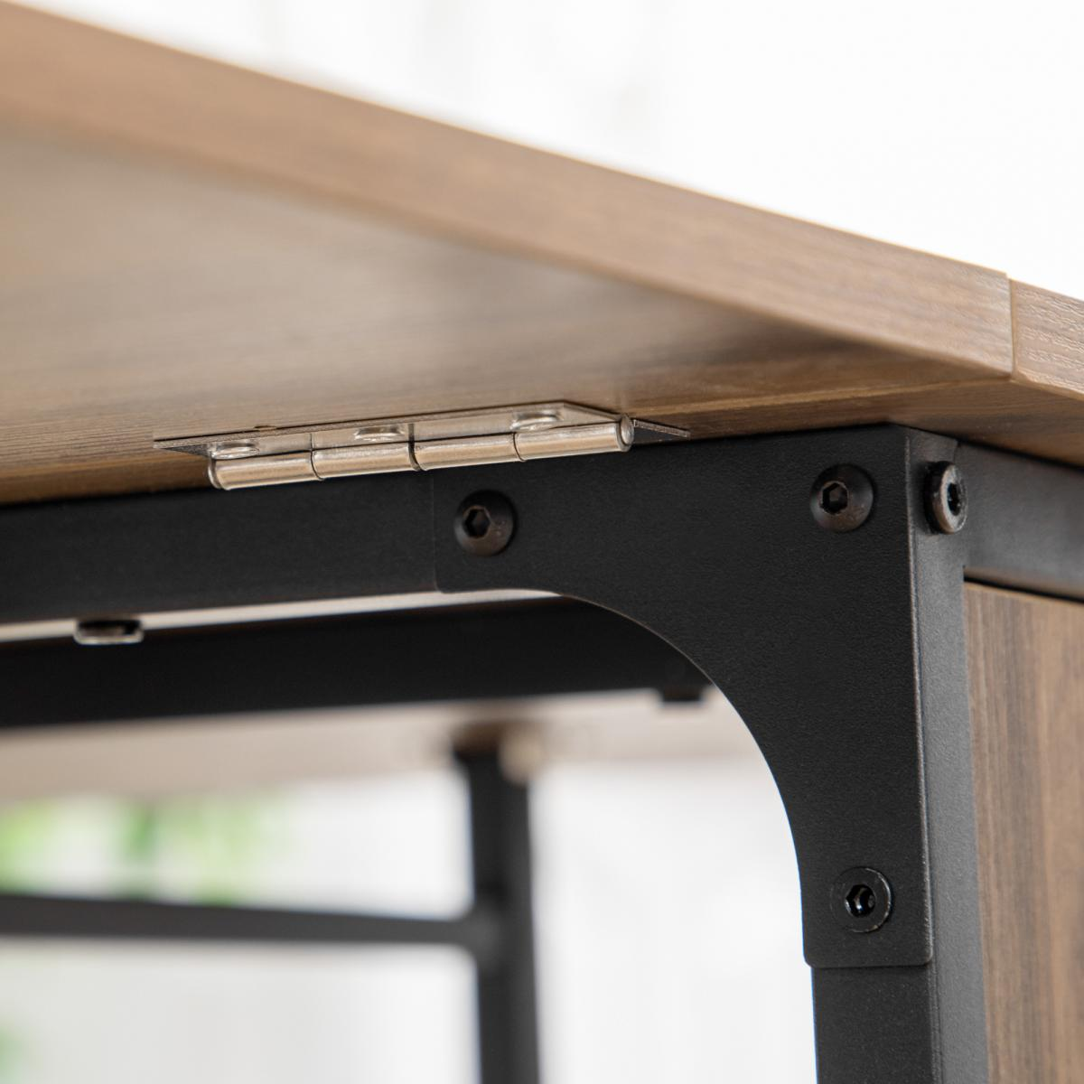 Nahaufnahme einer Tischkante. Der Holztisch hat ein Metallscharnier und einen schwarzen Metallrahmen. Schrauben sichern den Rahmen. Hintergrund ist unscharf.
