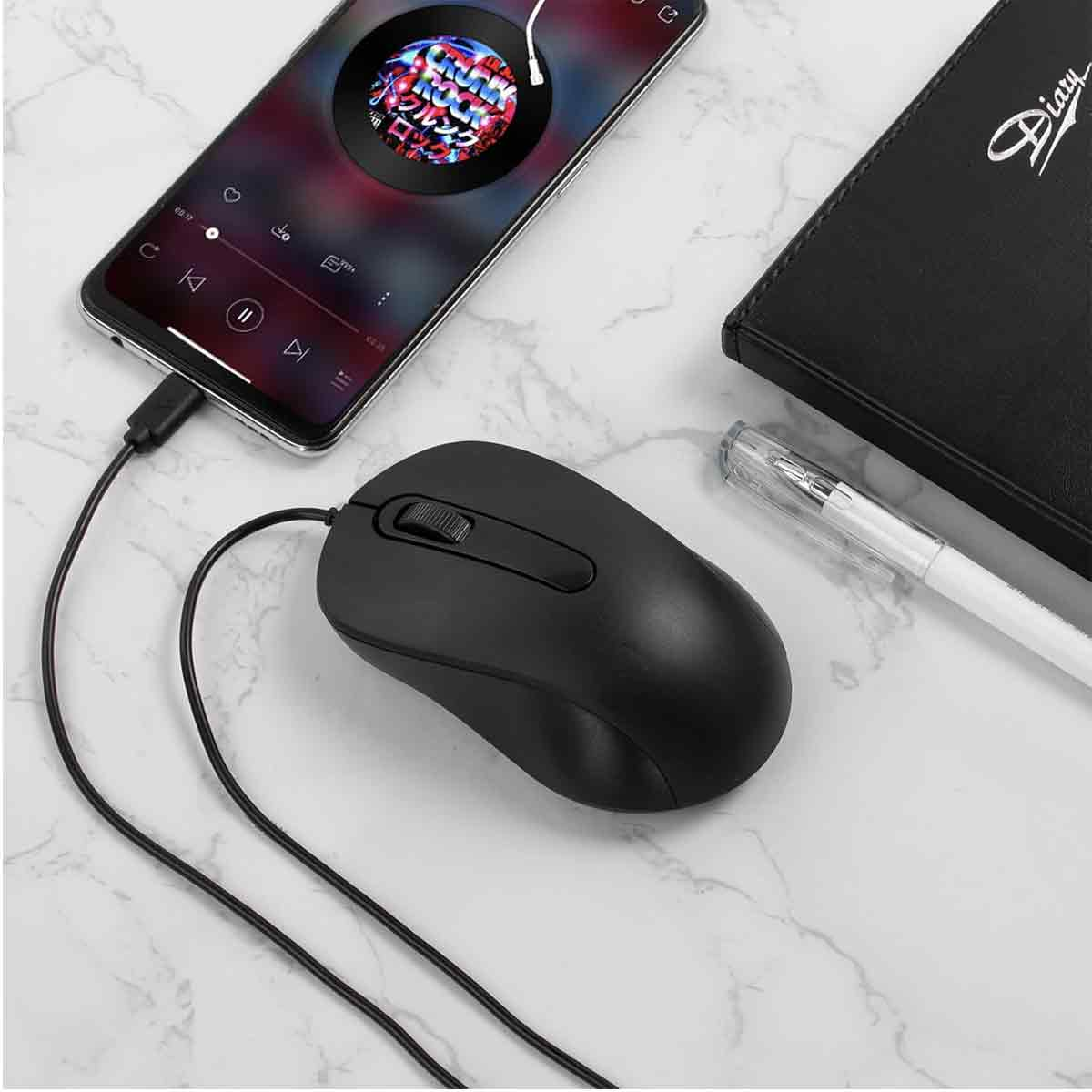 Eine schwarze Computermaus, ein Smartphone, ein schwarzes Tagebuch und ein Stift liegen auf einer Marmoroberfläche. Das Telefon spielt Musik.