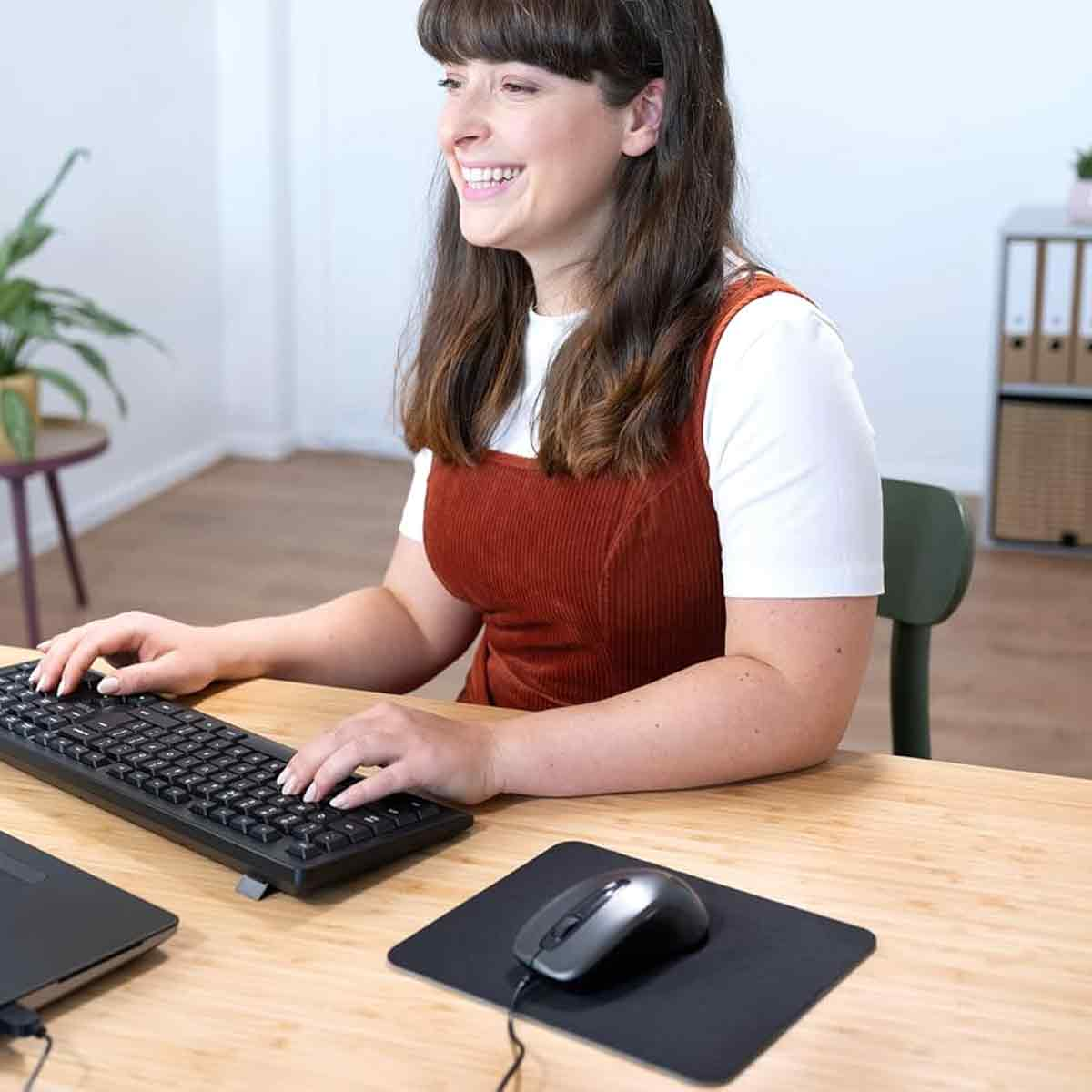 Eine Frau lächelt, während sie an einem Schreibtisch arbeitet. Sie tippt auf einer Tastatur mit Maus und Laptop. Ein Stuhl und ein Schrank sind im Hintergrund.