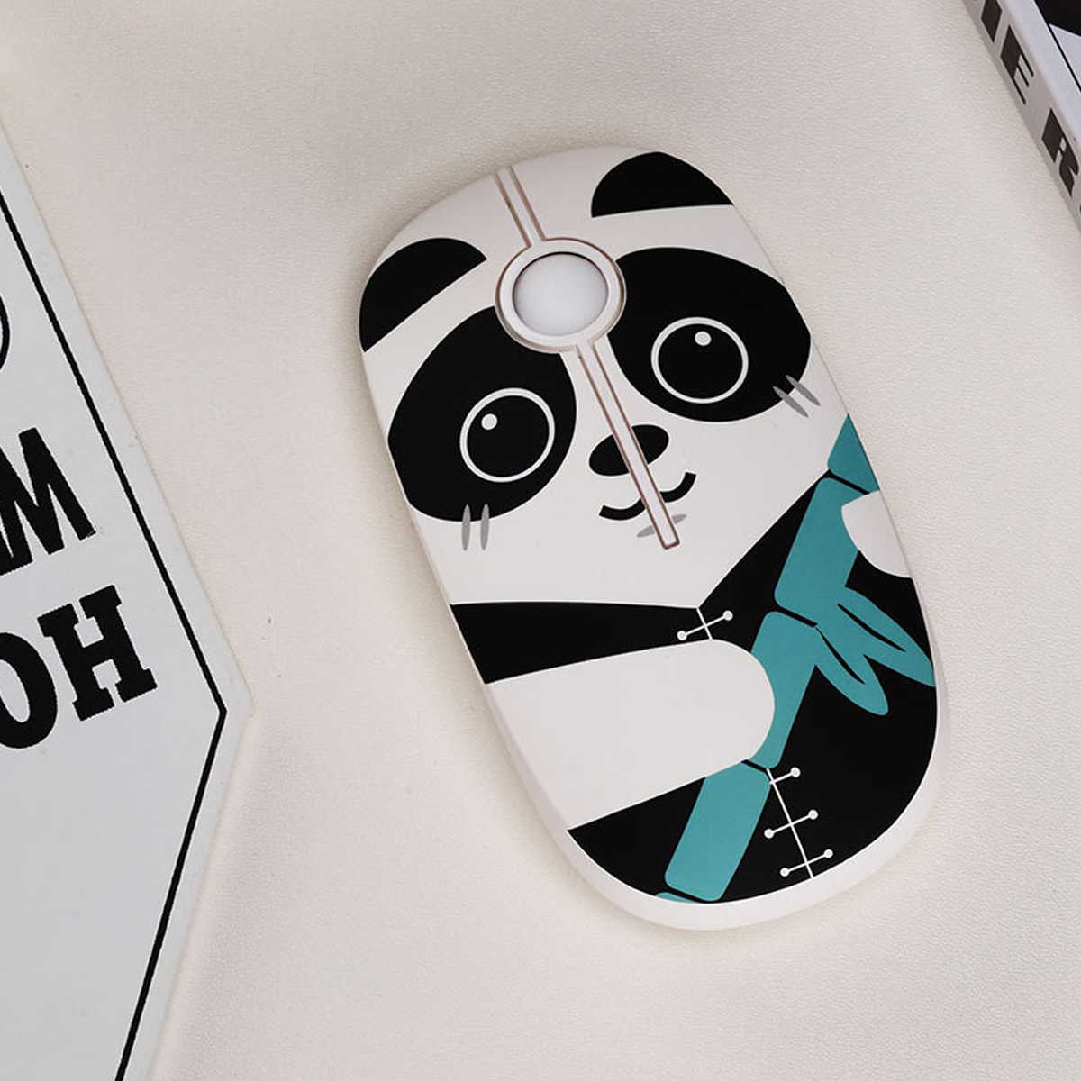 Eine weiße Maus mit Panda-Design. Sie hat schwarz-weiße Panda-Gesichtsdetails. Bambusblätter sind an der Seite. Sie liegt auf einer weißen Oberfläche.