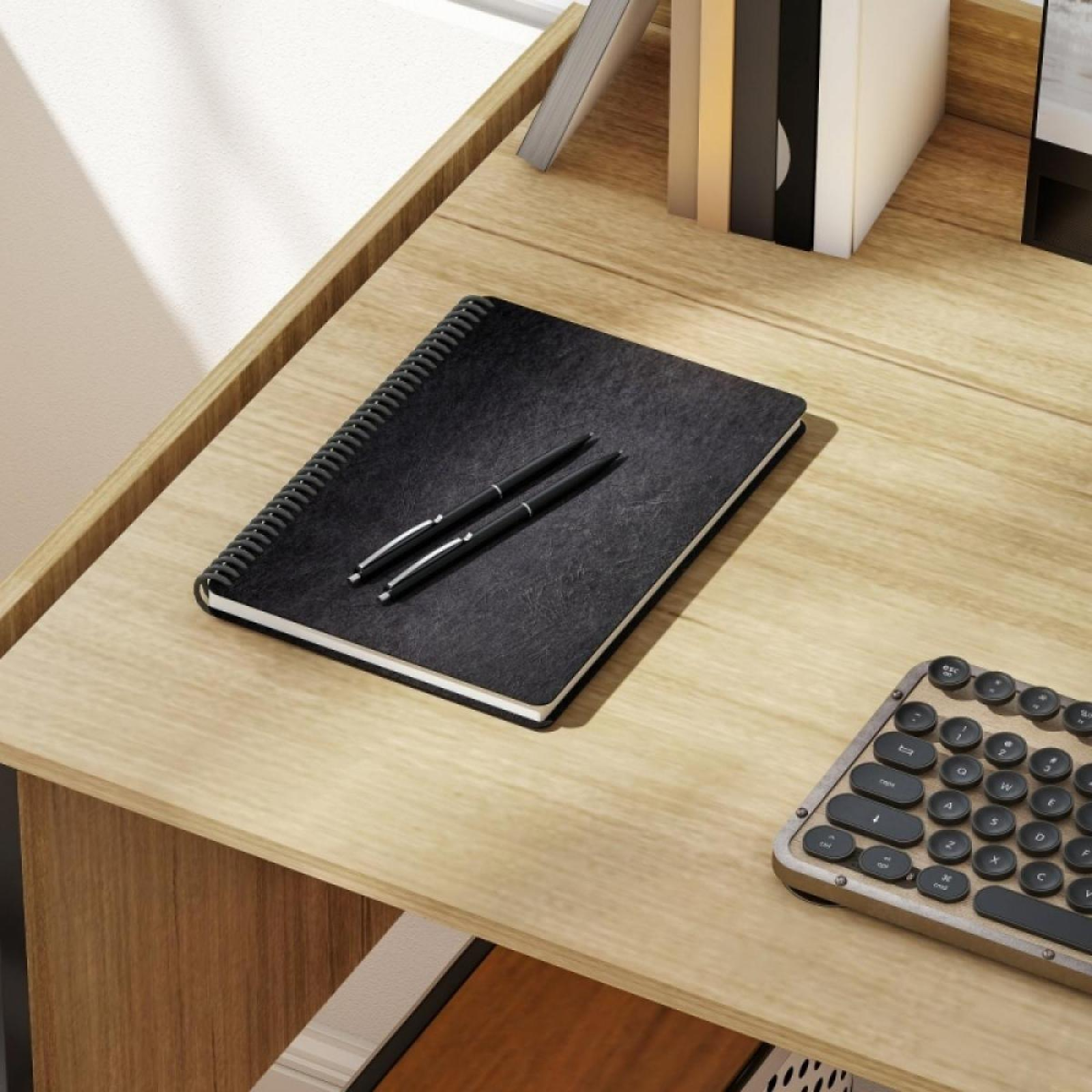 Ein schwarzes Notizbuch mit zwei Stiften liegt auf einem Holztisch. Eine Tastatur und Bücher sind ebenfalls zu sehen.