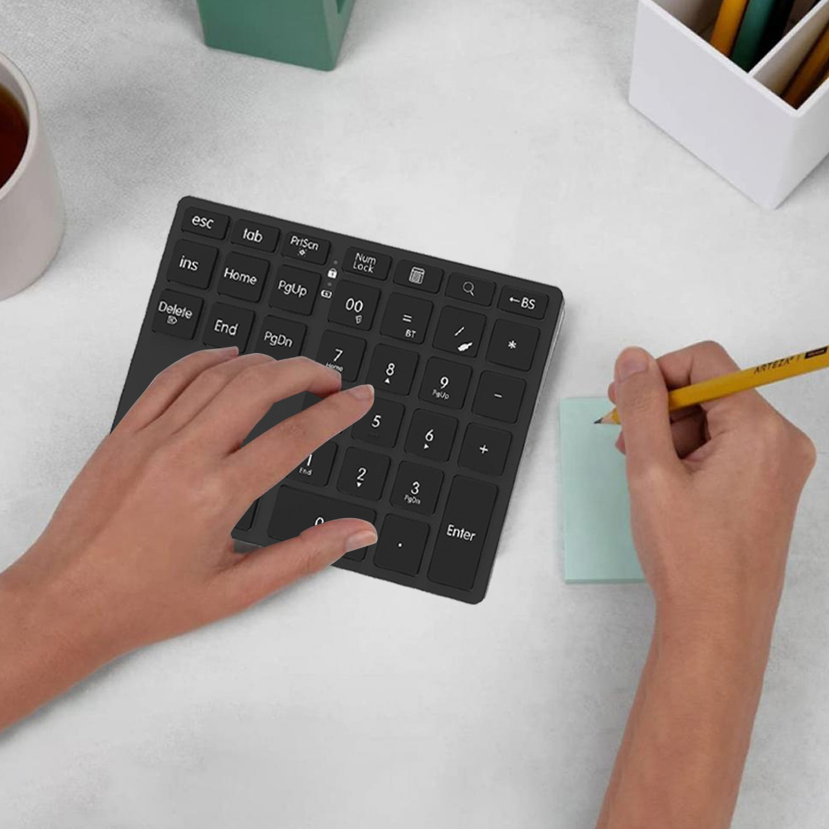 Hände benutzen eine schwarze Tastatur und schreiben mit einem gelben Bleistift auf einem Haftzettel auf einem weißen Schreibtisch.