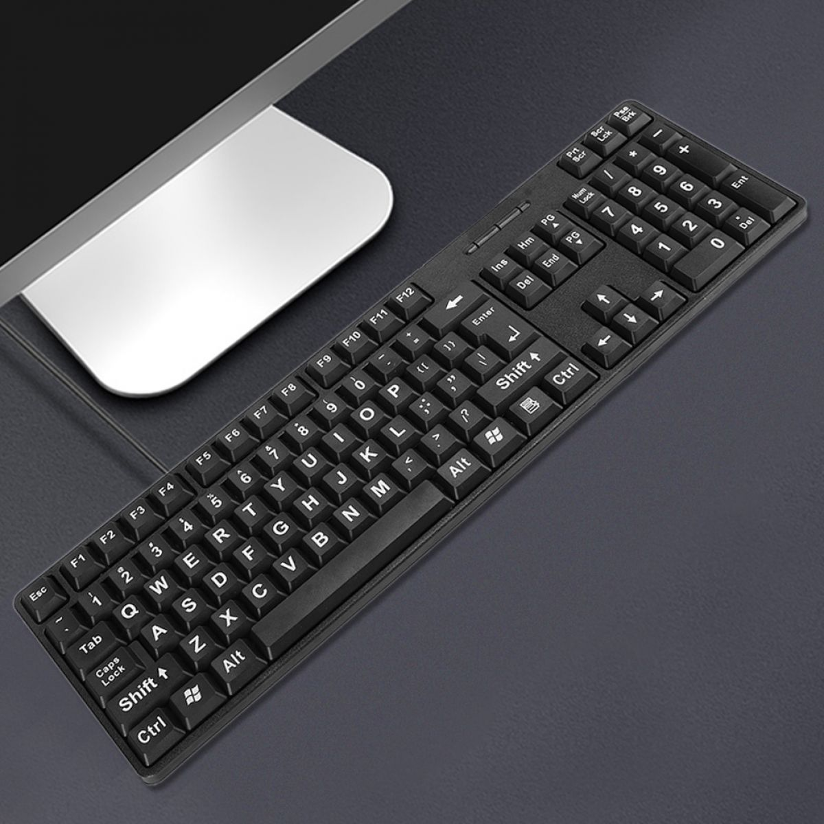 Schwarze Tastatur mit weißen Tasten, ein Computermonitor und ein dunkler Schreibtisch. Die Tastatur ist zum Betrachter abgewinkelt.