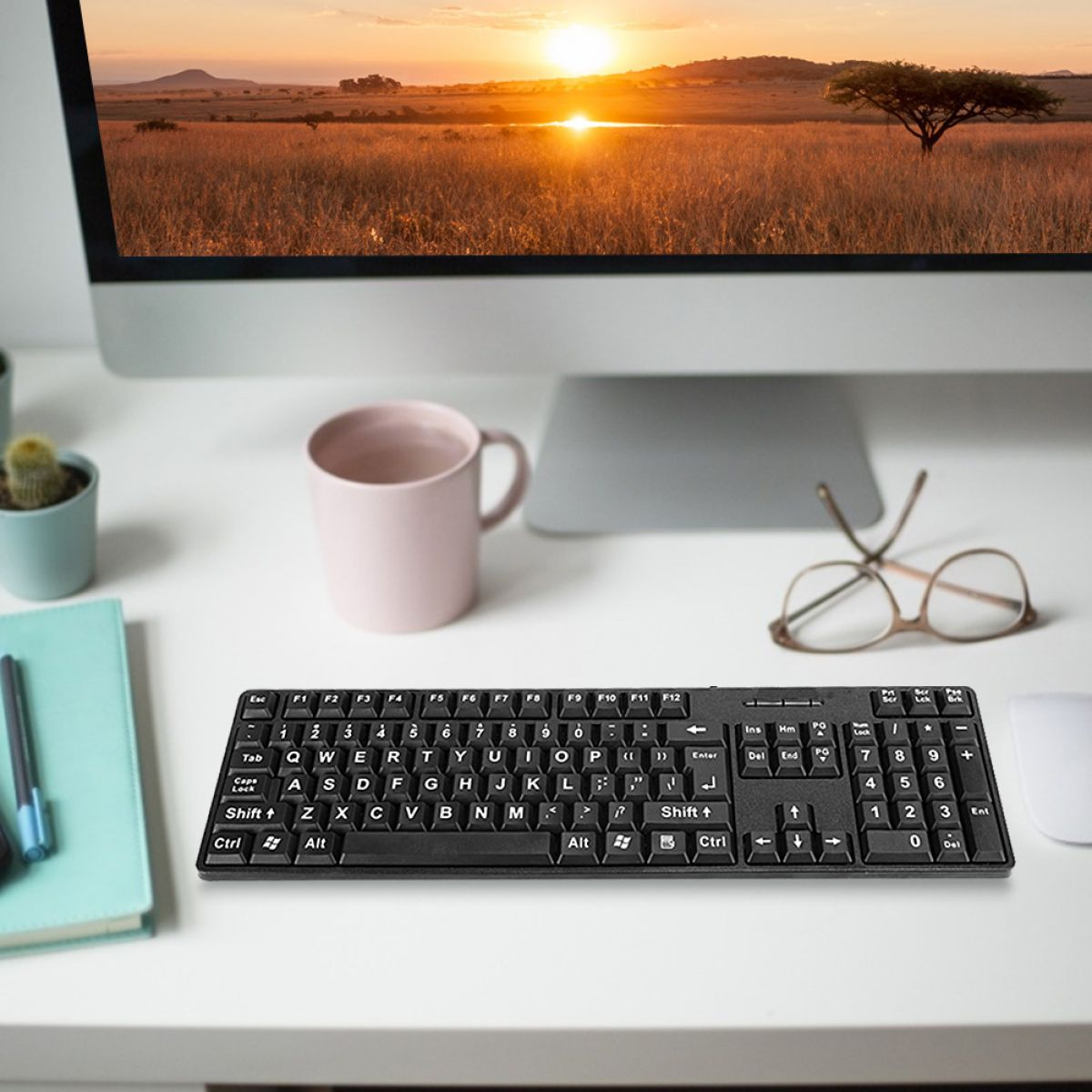 Eine schwarze Tastatur, ein rosa Becher, eine Brille und ein Computermonitor mit Sonnenuntergang über einer Savanne stehen auf einem weißen Schreibtisch.