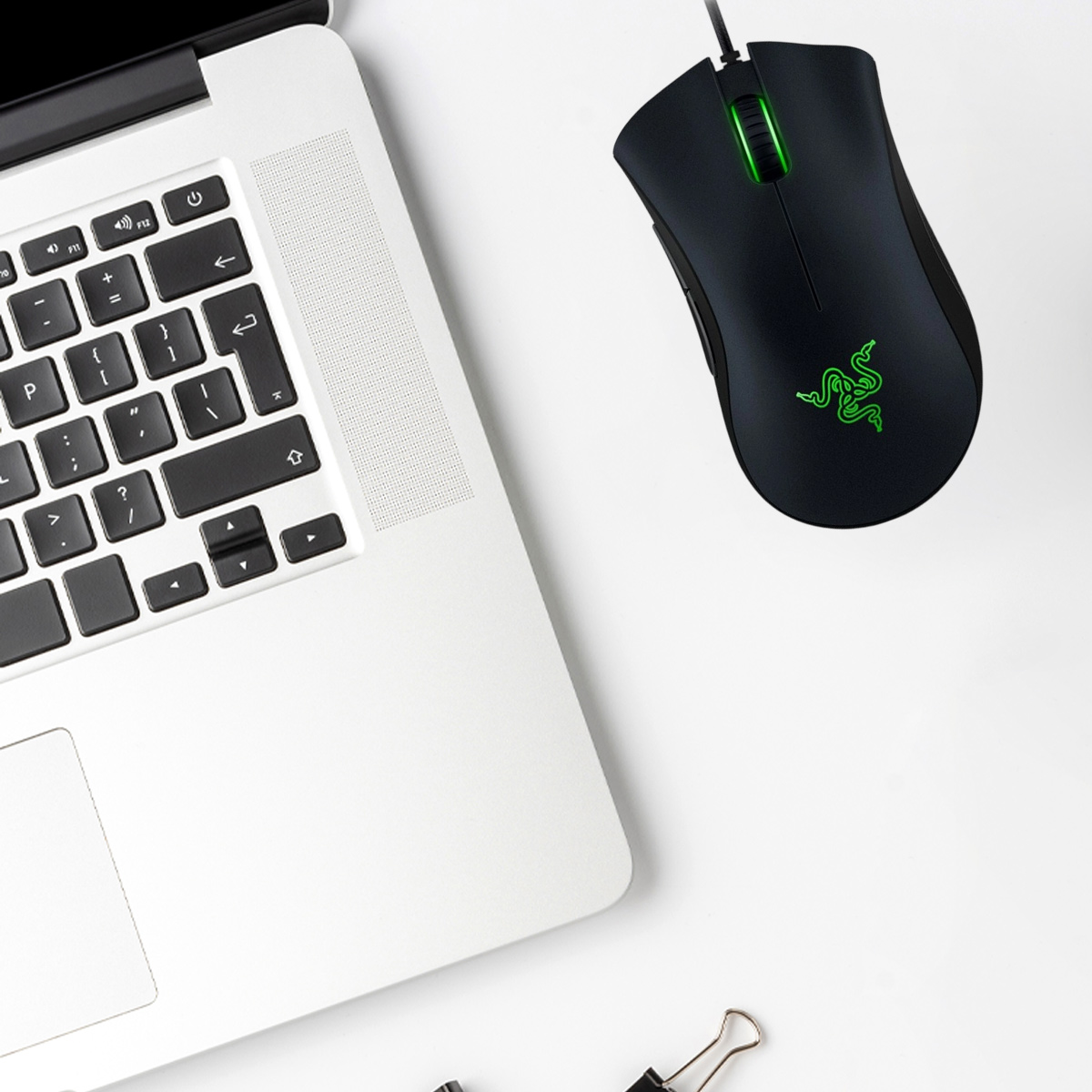Ein Laptop und eine schwarze Razer-Maus mit grünem Logo und Akzenten liegen auf einer weißen Oberfläche. Am Boden befindet sich eine Büroklammer.
