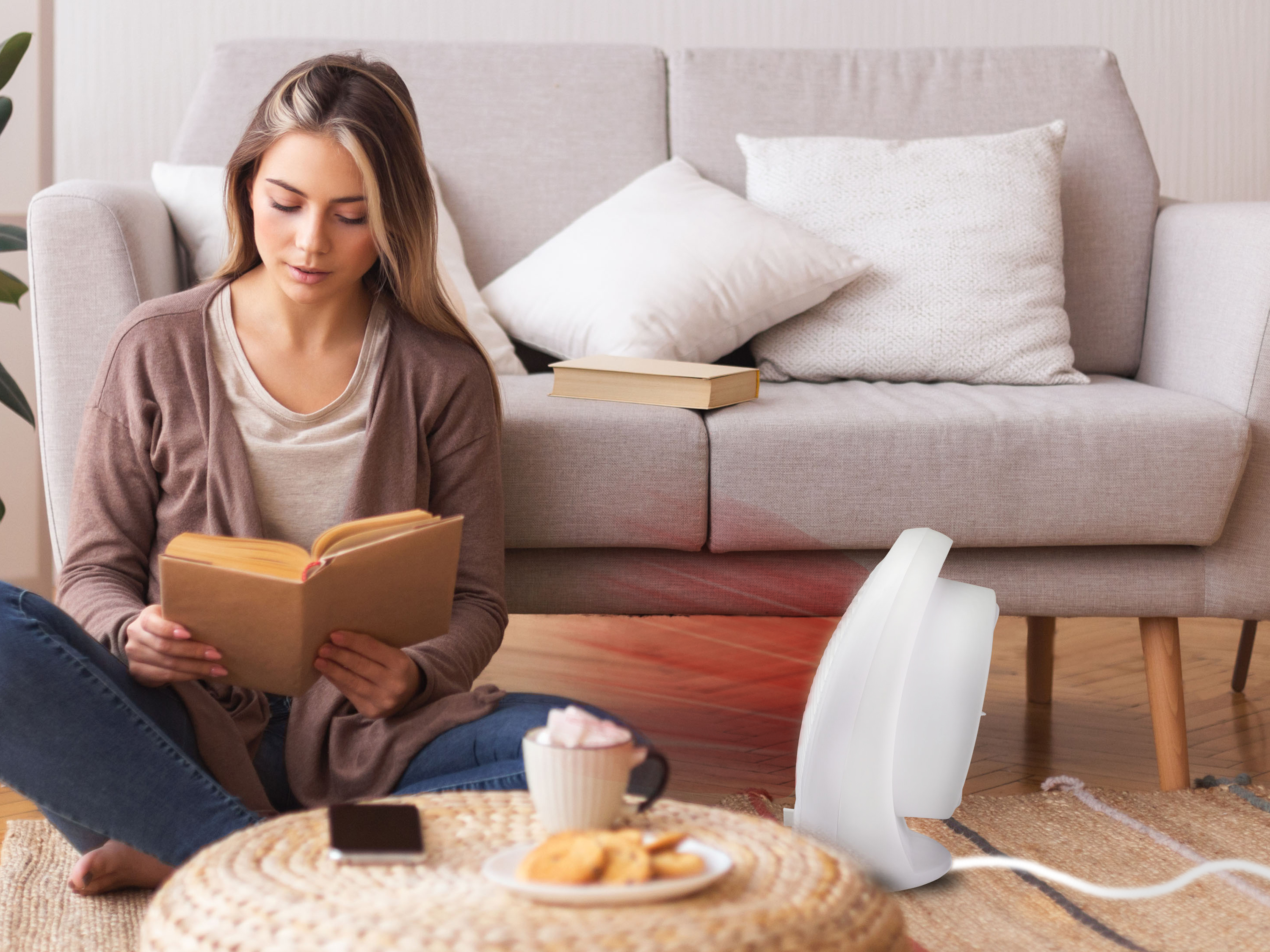 Donna che legge, seduta vicino a un divano, caffè, biscotti e un riscaldatore. Un libro sul divano.