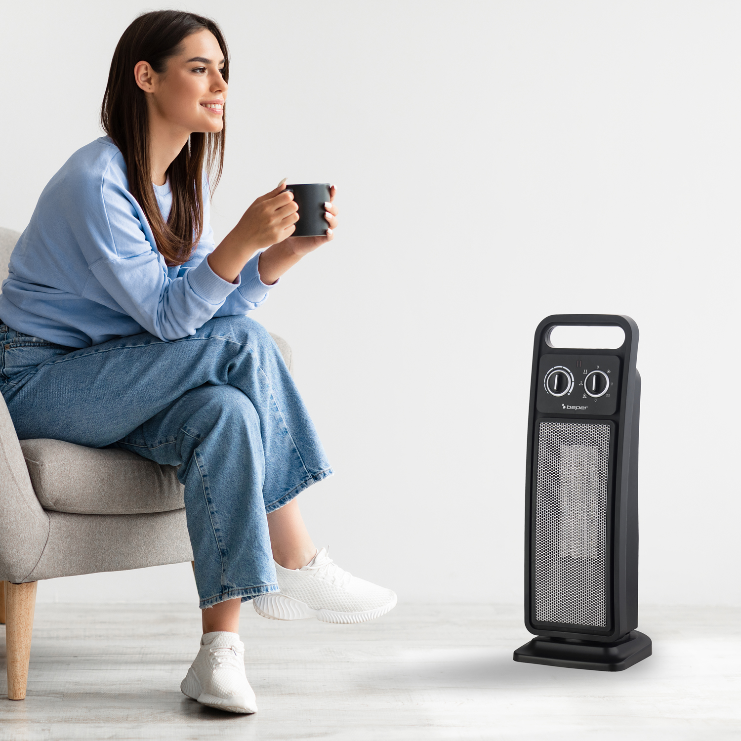 Una donna in blu tiene una tazza. Un termoventilatore nero è accanto a lei. È in una stanza luminosa.