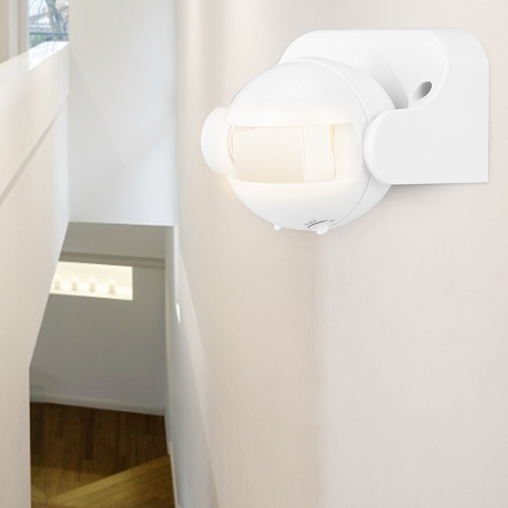 Ein weißer Bewegungsmelder ist an einer weißen Wand befestigt. Eine Treppe führt hinab, mit sichtbaren Holzstufen. Sonnenlicht scheint aus einem Fenster.