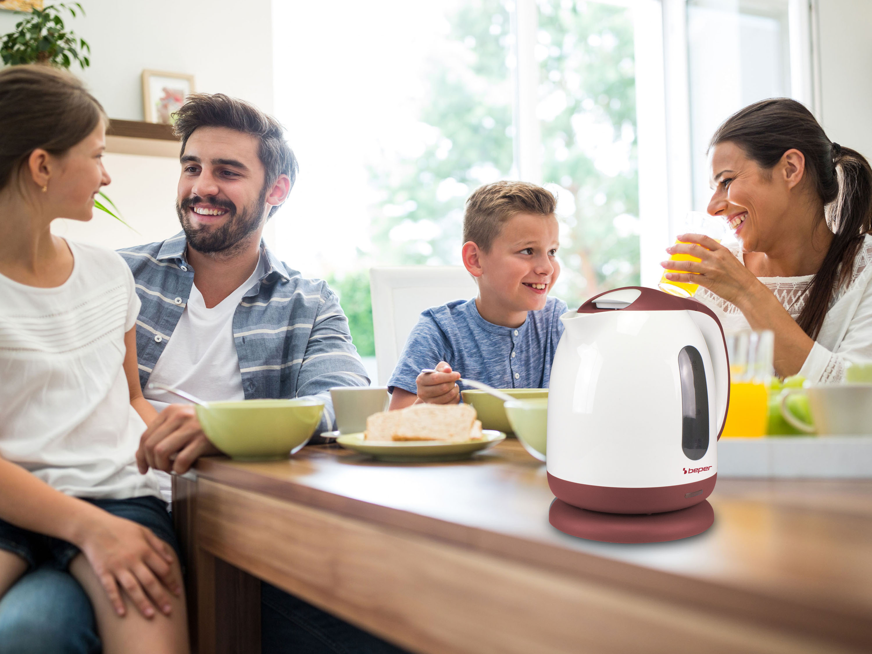Una famiglia fa colazione. Un bollitore bianco e marrone è sul tavolo. Tutti sorridono e ci sono piatti di cibo.