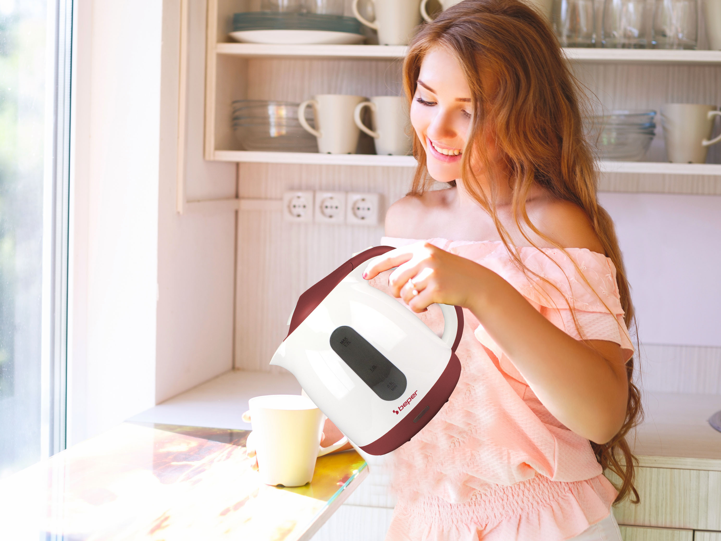 Una donna con un top rosa versa da un bollitore bianco e bordeaux in una tazza. Mensole della cucina con piatti sullo sfondo.
