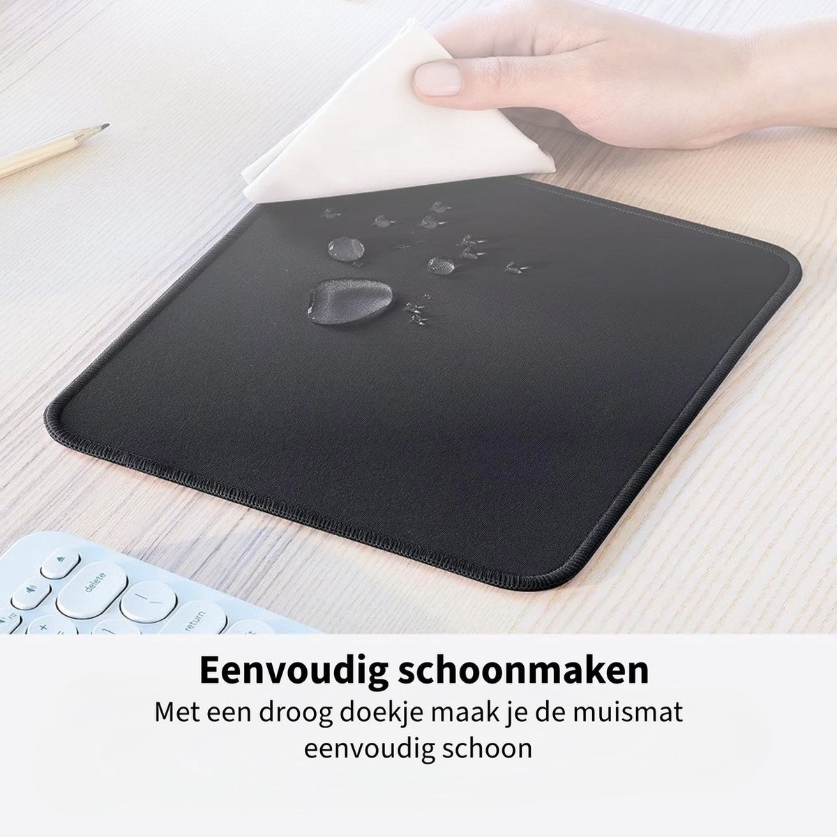 A hand wipes a black mousepad with a white cloth. Water droplets are visible. Keyboard and pencil are on the light-colored desk.