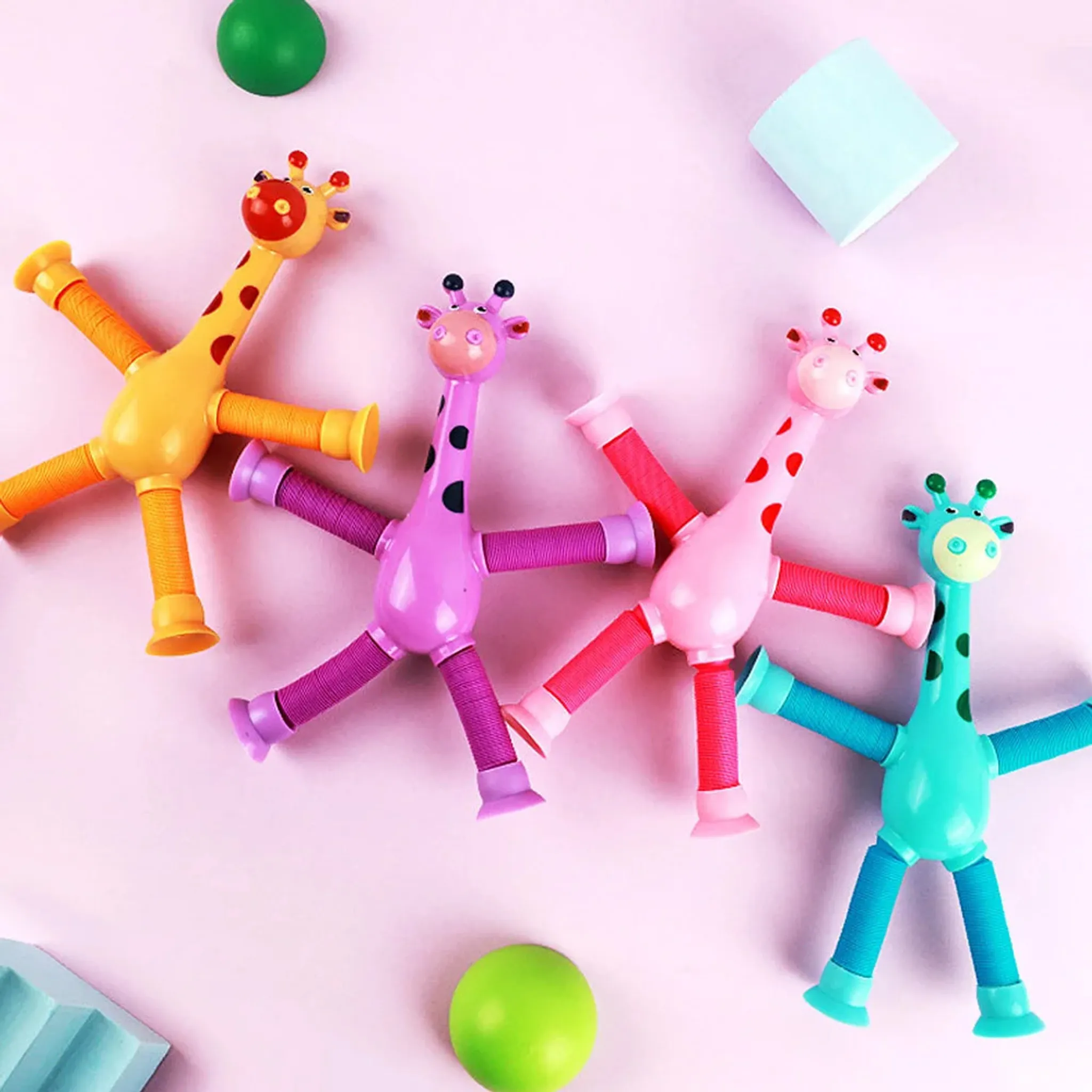 Vier bunte Giraffenspielzeuge mit ausziehbaren Gliedmaßen auf rosa Hintergrund, zusammen mit zwei grünen Bällen und einem blauen Block.