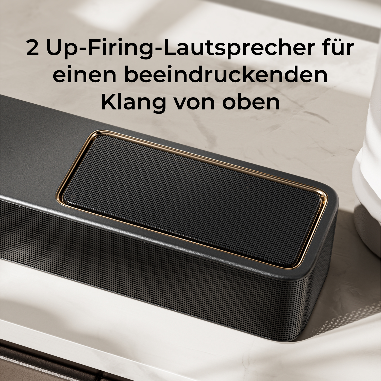 Eine schwarze Soundbar mit goldenem Rand liegt auf einer weißen Oberfläche. Oben steht: '2 Up-Firing-Lautsprecher...'