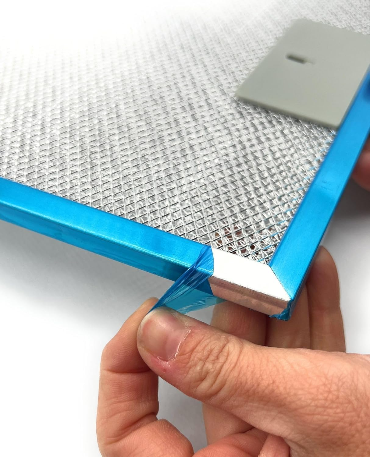 Una mano despega una película protectora azul de un filtro metálico de campana extractora. Una herramienta cuadrada gris descansa cerca de la malla.