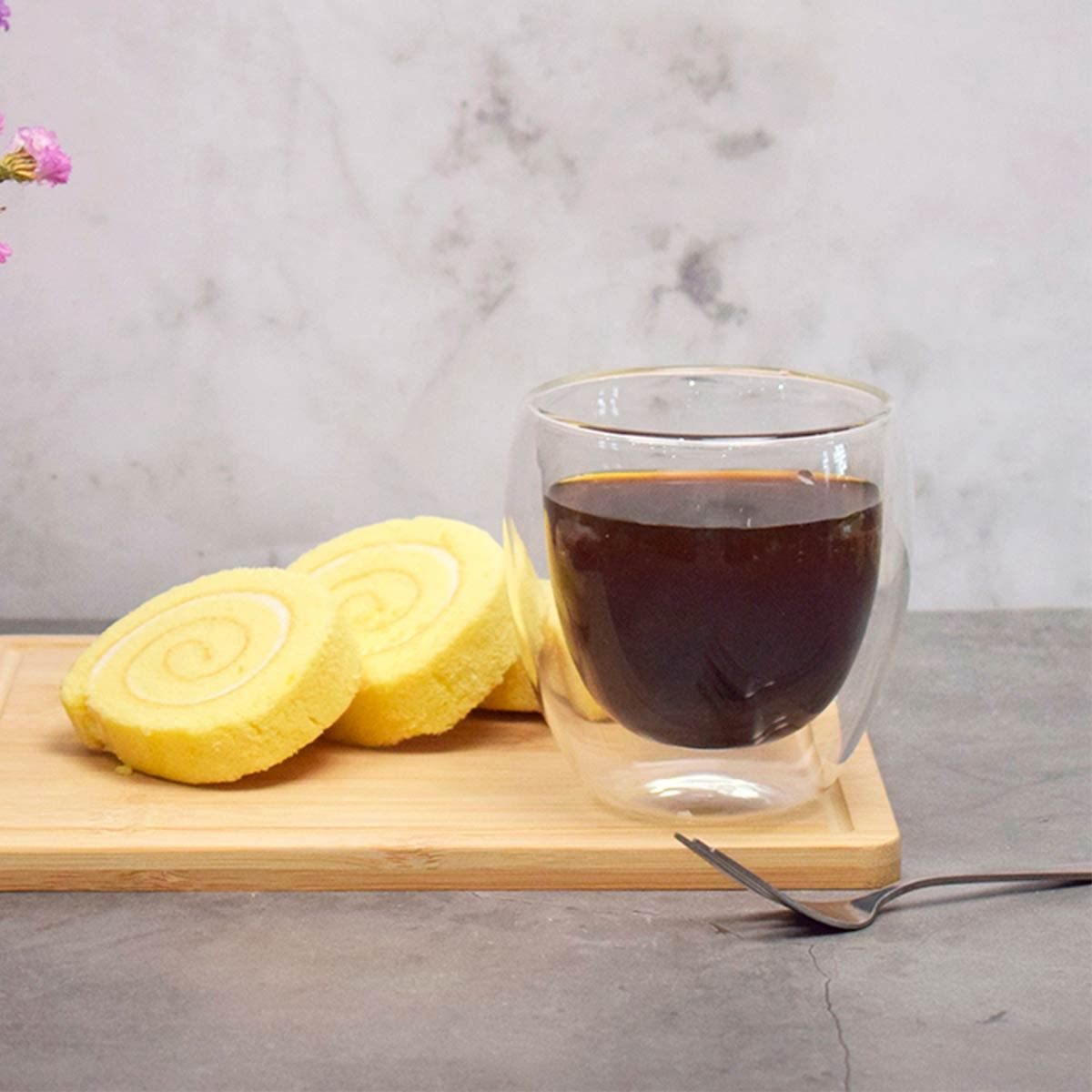 Café en vaso, rebanadas de bizcocho enrollado en tabla de madera, tenedor y flores sobre superficie gris.