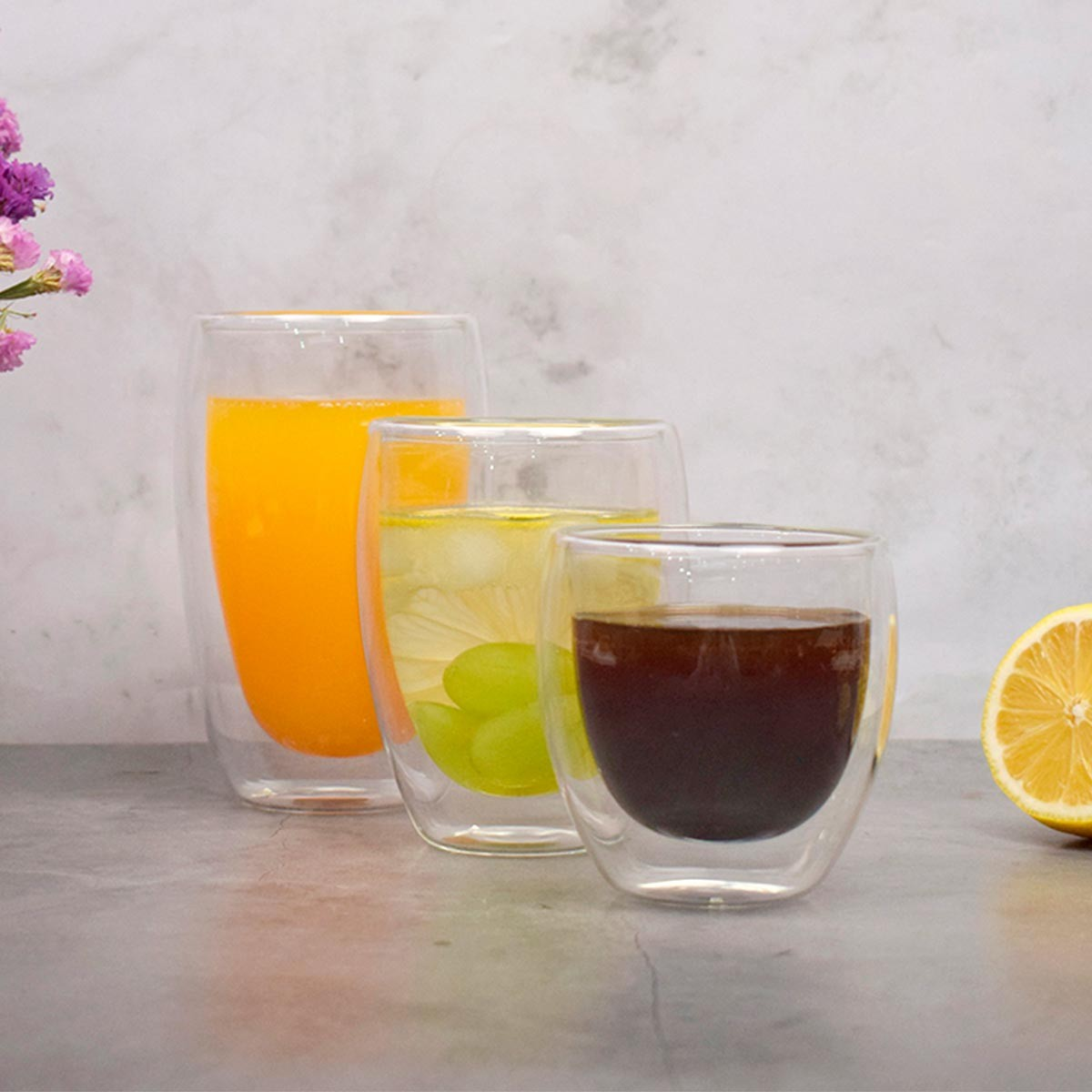 Tres vasos transparentes con diferentes bebidas: jugo de naranja, un cóctel con hielo y uvas, y una bebida oscura. Media rodaja de limón visible.