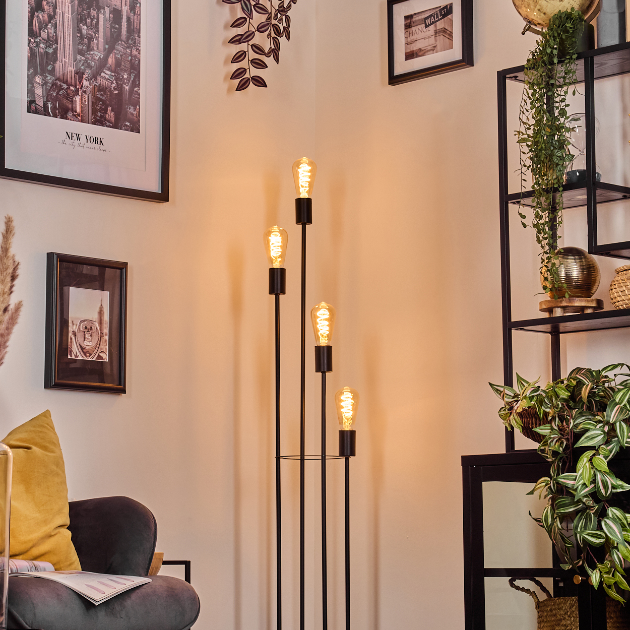 Eine Stehlampe mit vier Edison-Glühbirnen steht in einem Raum mit Bildern und Pflanzen. Links ist ein grauer Stuhl.