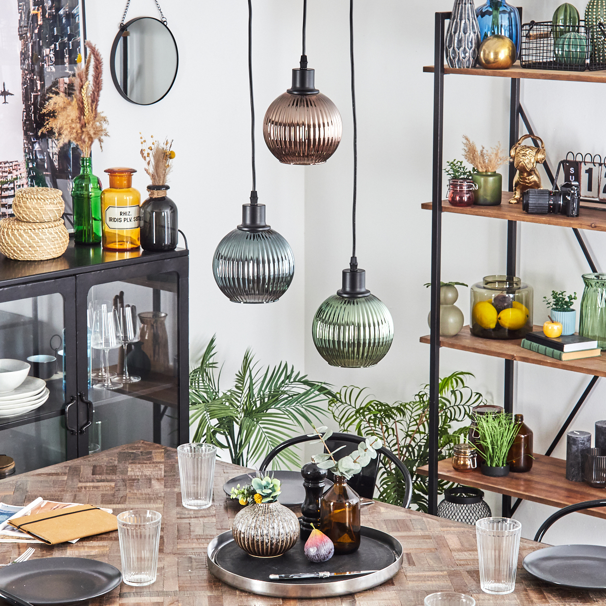Esszimmer. Drei Pendelleuchten hängen über einem Holztisch, der mit Tellern und Gläsern gedeckt ist. Regale mit Dekoration im Hintergrund.