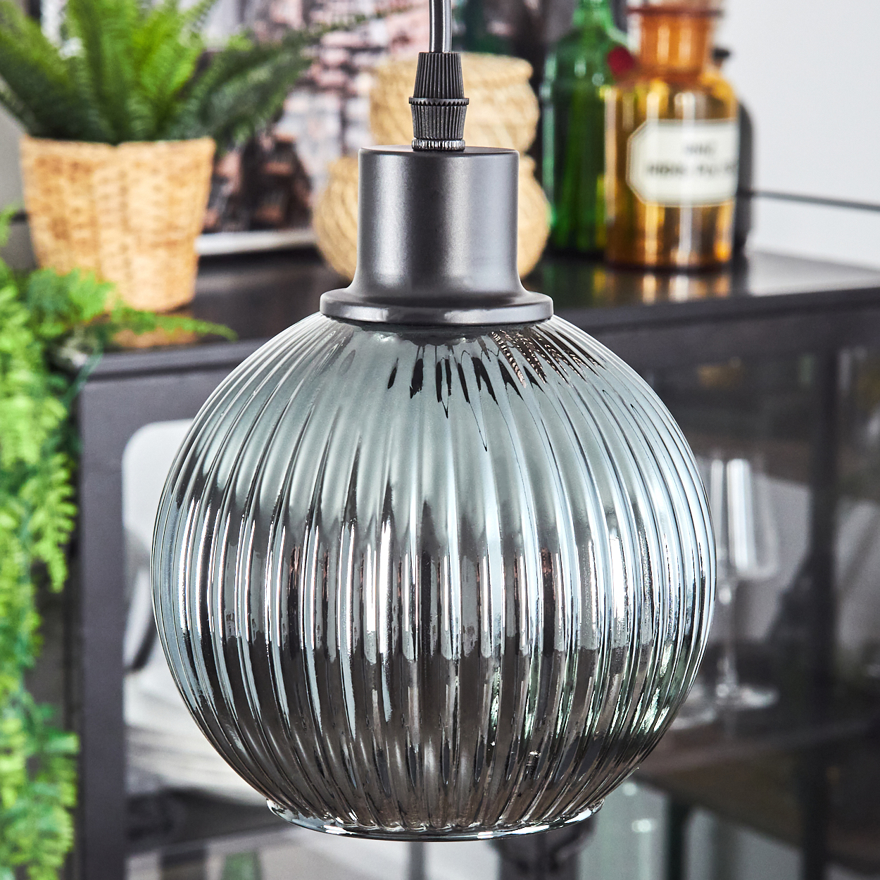 Eine graue, gerippte Glashängelampe hängt. Ein Topf mit Pflanze und Dekoration sind im Hintergrund. Die Lampe hat eine schwarze Halterung.