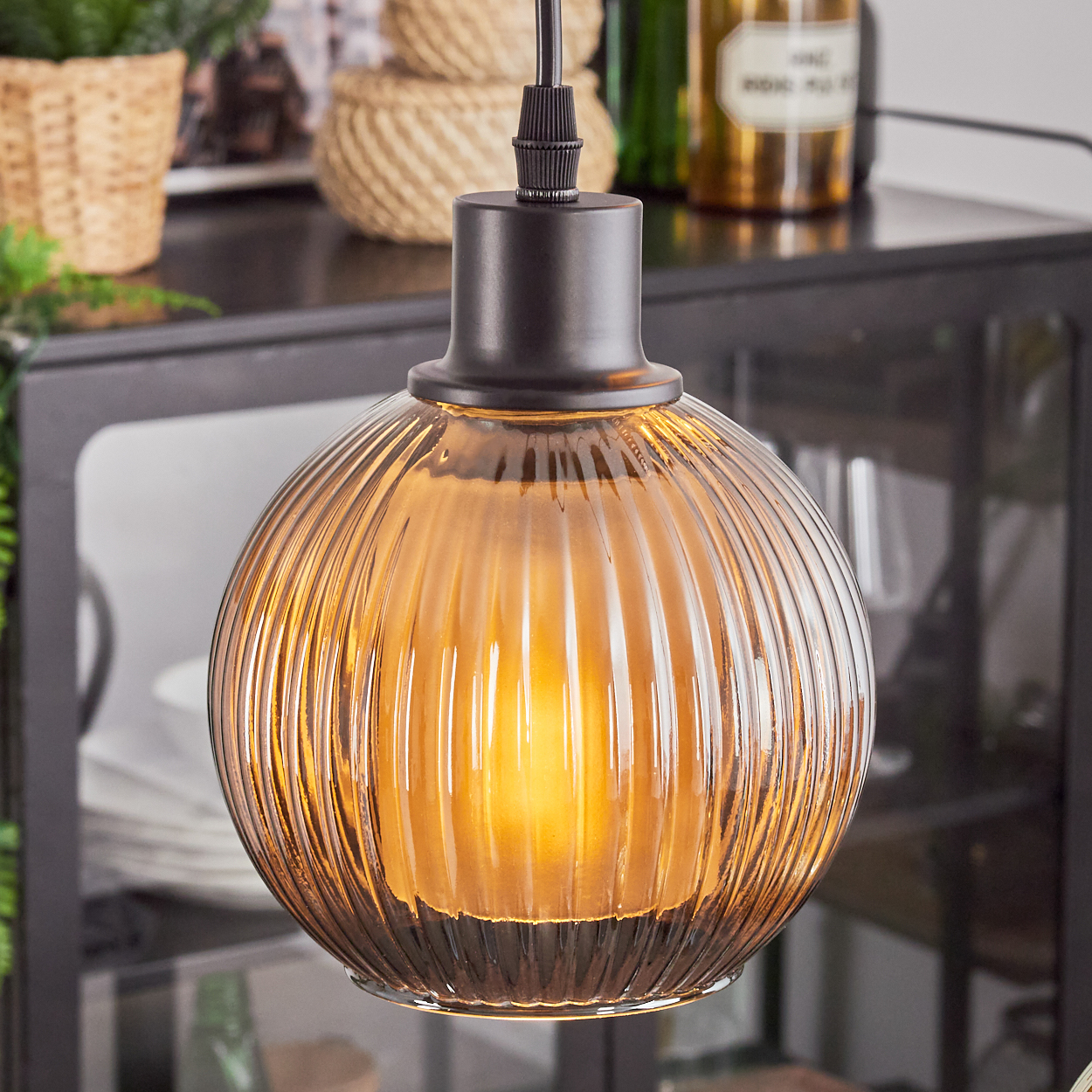 Eine Glas-Pendelleuchte mit geripptem Design. Sie hat einen warmen, bernsteinfarbenen Schein. Der Hintergrund umfasst Dekorationsartikel und einen dunklen Schrank.