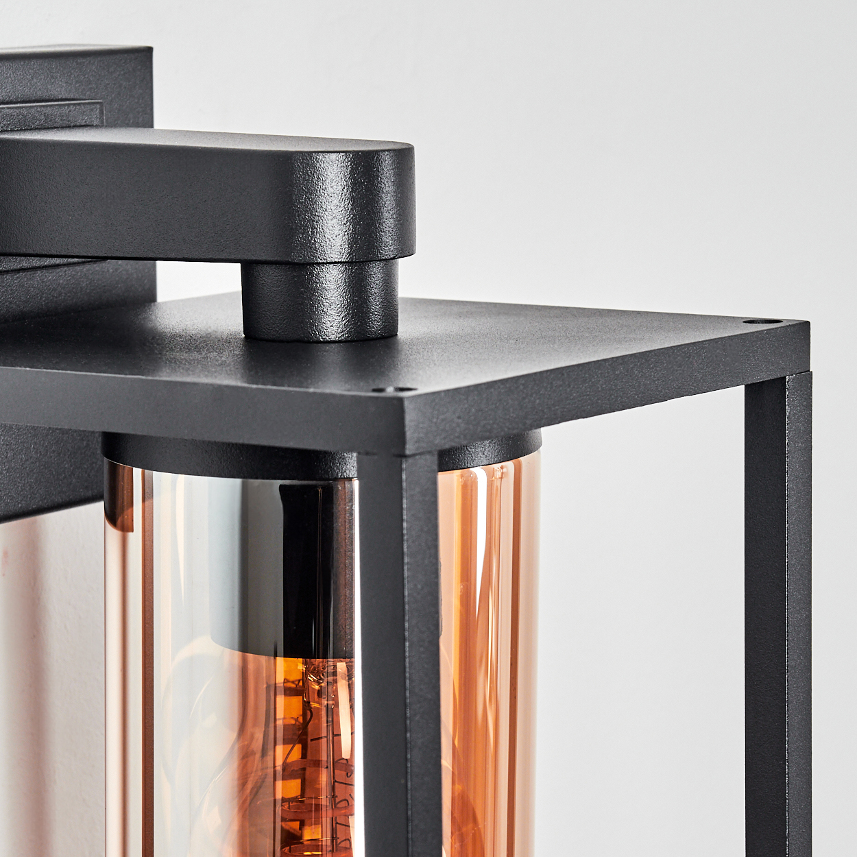 Nahaufnahme einer schwarzen Außenlampe. Sie hat einen rechteckigen Rahmen, einen zylindrischen Glasschirm und eine warmtonige Glühbirne.