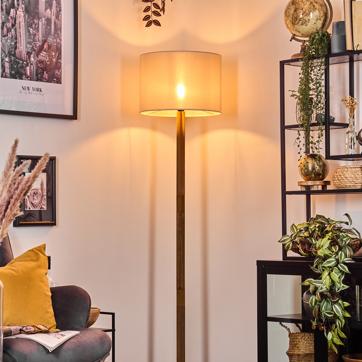 Eine beleuchtete Stehlampe steht in einem Raum. Bücherregale, ein Sofa mit gelbem Kissen und gerahmte Kunst sind im Hintergrund sichtbar.