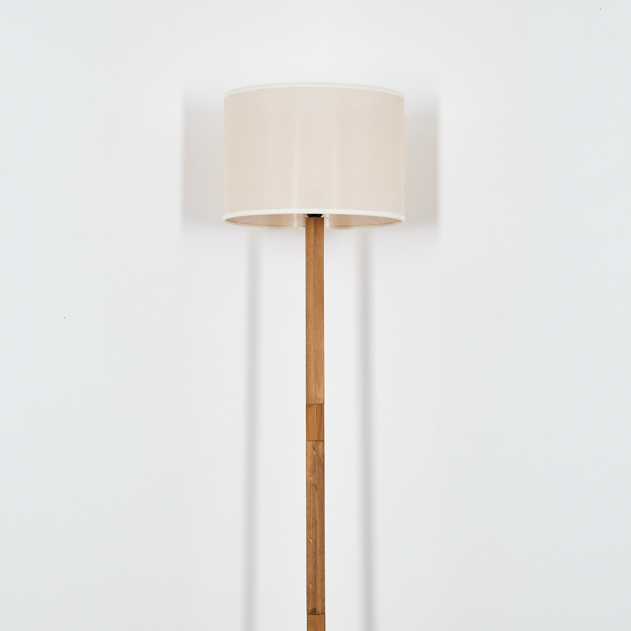 Ein beigefarbener Lampenschirm sitzt auf einer Holzstange. Der Hintergrund ist eine schlichte weiße Wand. Die Lampe wirft einen Schatten.