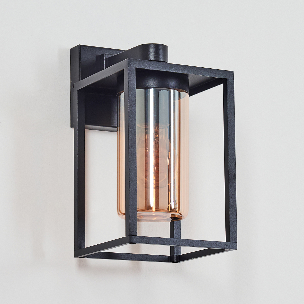 Schwarze Metall-Außenleuchte an einer weißen Wand. Amberfarbener Glaszylinder in einem schwarzen Rahmen mit Glühbirne.