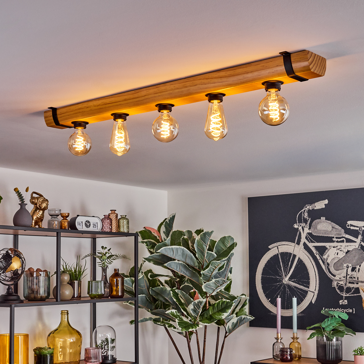 Eine Deckenleuchte mit Holzbalken und fünf Vintage-Glühbirnen. Darunter ein Regal mit Deko und Pflanzen. Motorrad-Kunstwerk an der Wand.