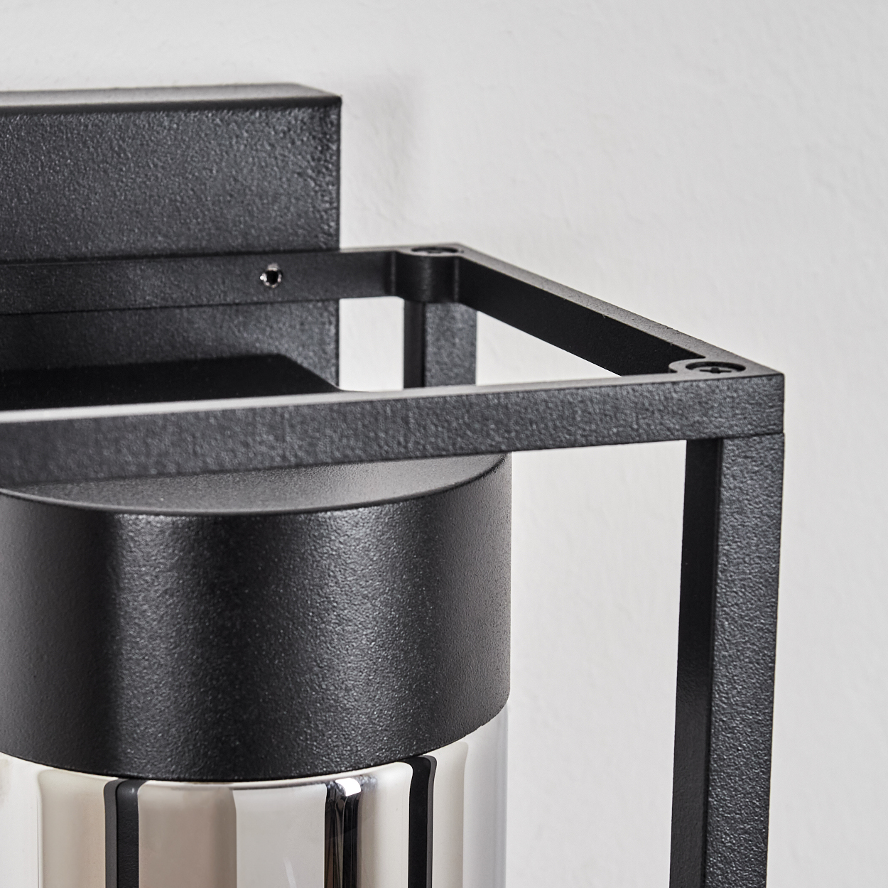 Nahaufnahme einer schwarzen Metall-Lampenhalterung. Sie hat einen quadratischen Rahmen und eine zylindrische Basis. Die Basis ist silber und reflektierend.