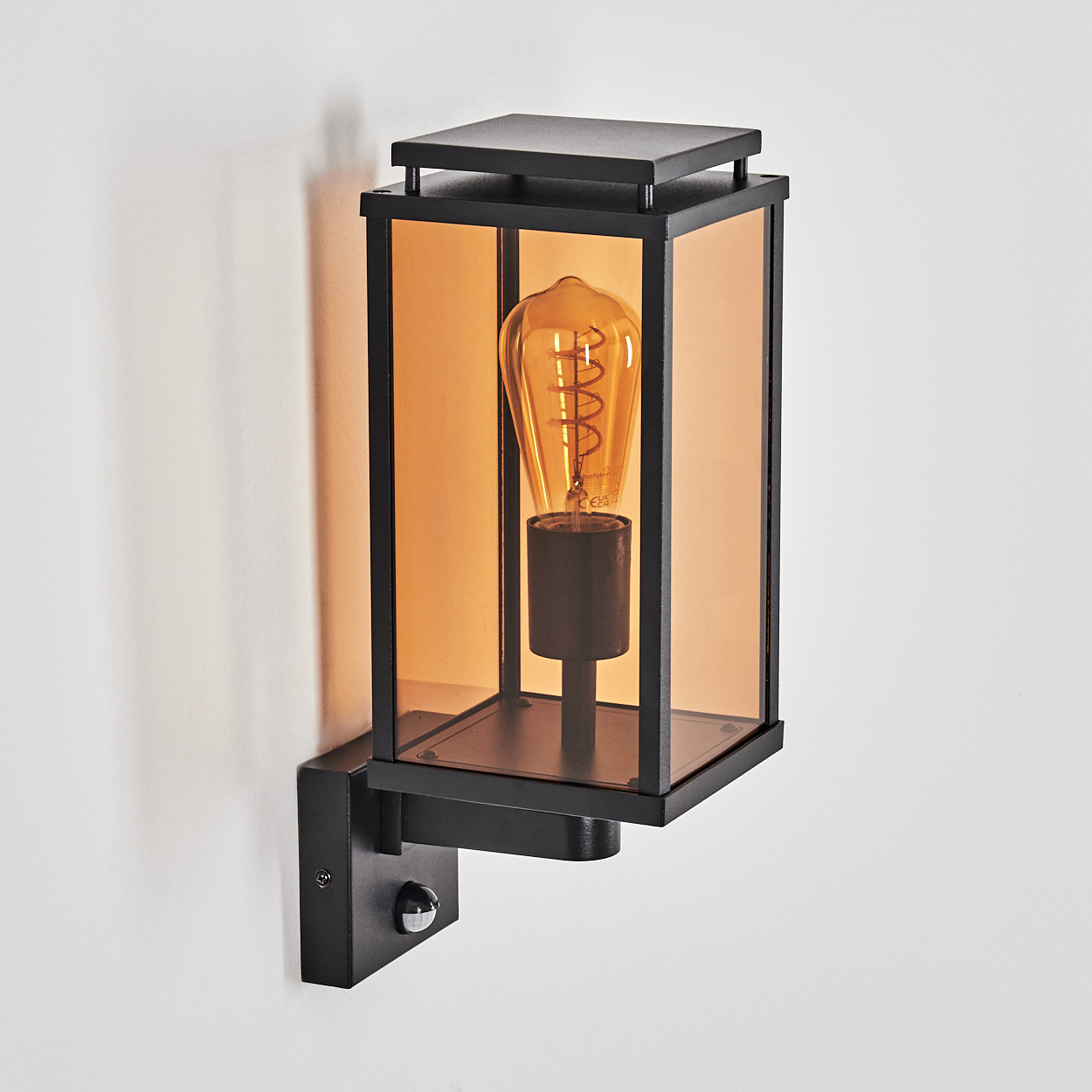 Schwarze Außenlampe an einer weißen Wand. Sie hat eine rechteckige Form mit bernsteinfarbenem Glas und einer sichtbaren Glühbirne.