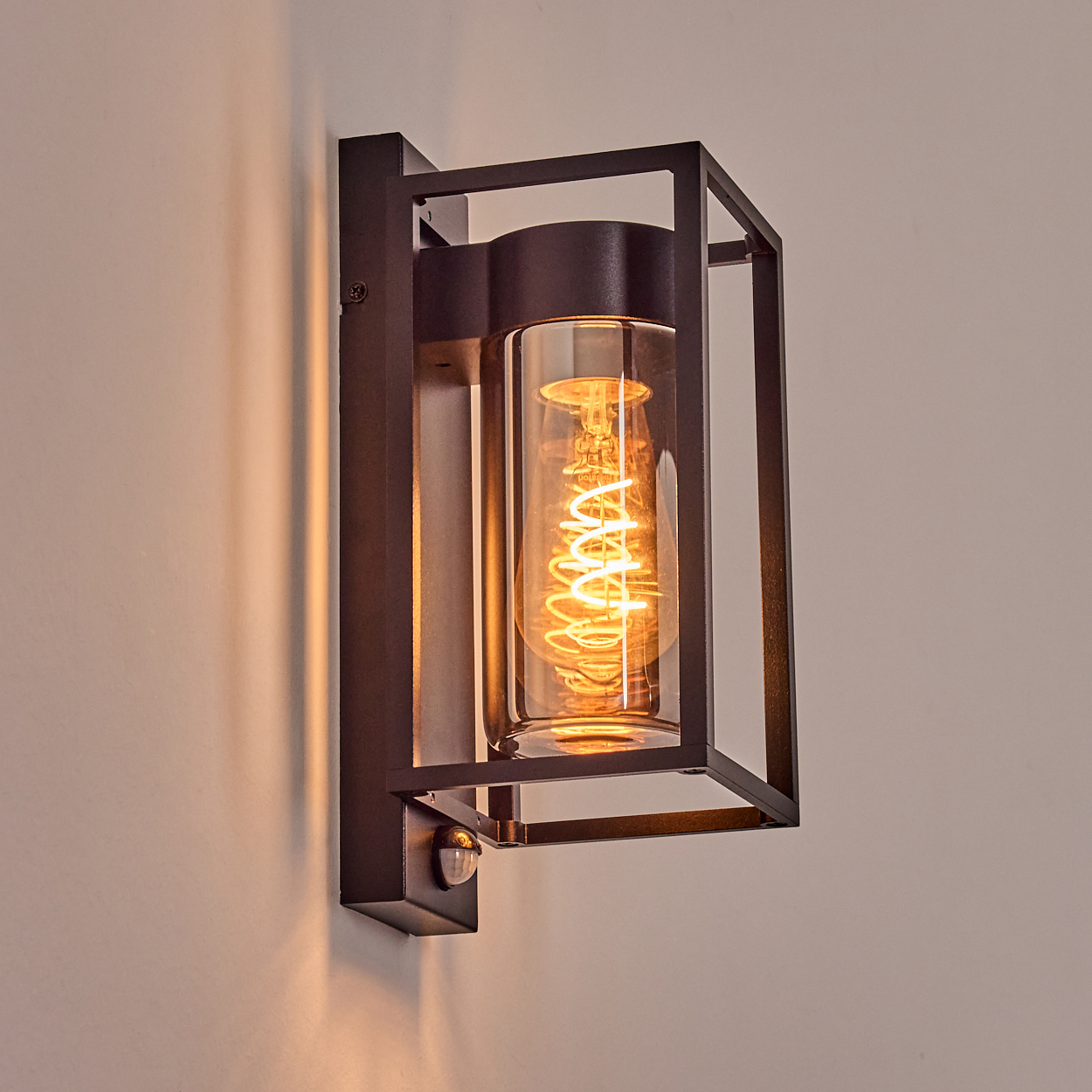 Eine schwarze Wandleuchte mit einem Glasrohr, das eine Wendelglühbirne enthält, ist an einer hellen Wand montiert. Das Licht wirft einen warmen Schein.