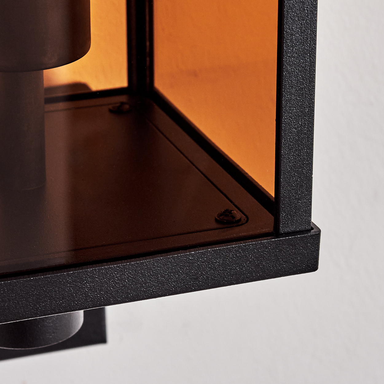 Nahaufnahme einer schwarzen Metalllaterne mit orangefarbenen Glasscheiben. Die Laterne ist an einer weißen Wand, Detail der Ecke sichtbar.