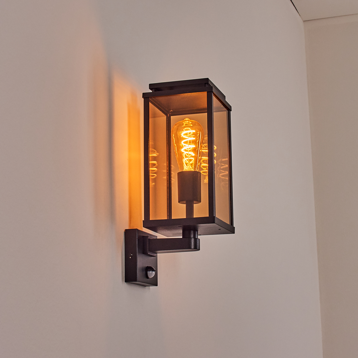 Eine schwarze Außenwandleuchte mit einer leuchtenden Edison-Glühbirne, an einer weißen Wand. Das Licht wirft einen orangen Schein.