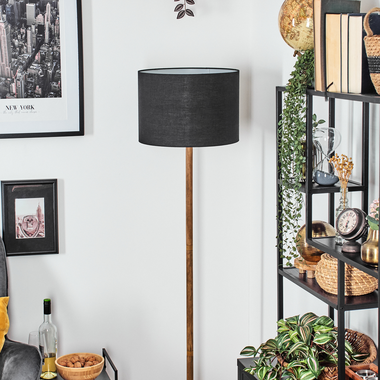 Eine Stehlampe mit schwarzem Lampenschirm steht in einem Raum mit einem Regal voller Dekoration. Ein Bild von New York hängt an der Wand.