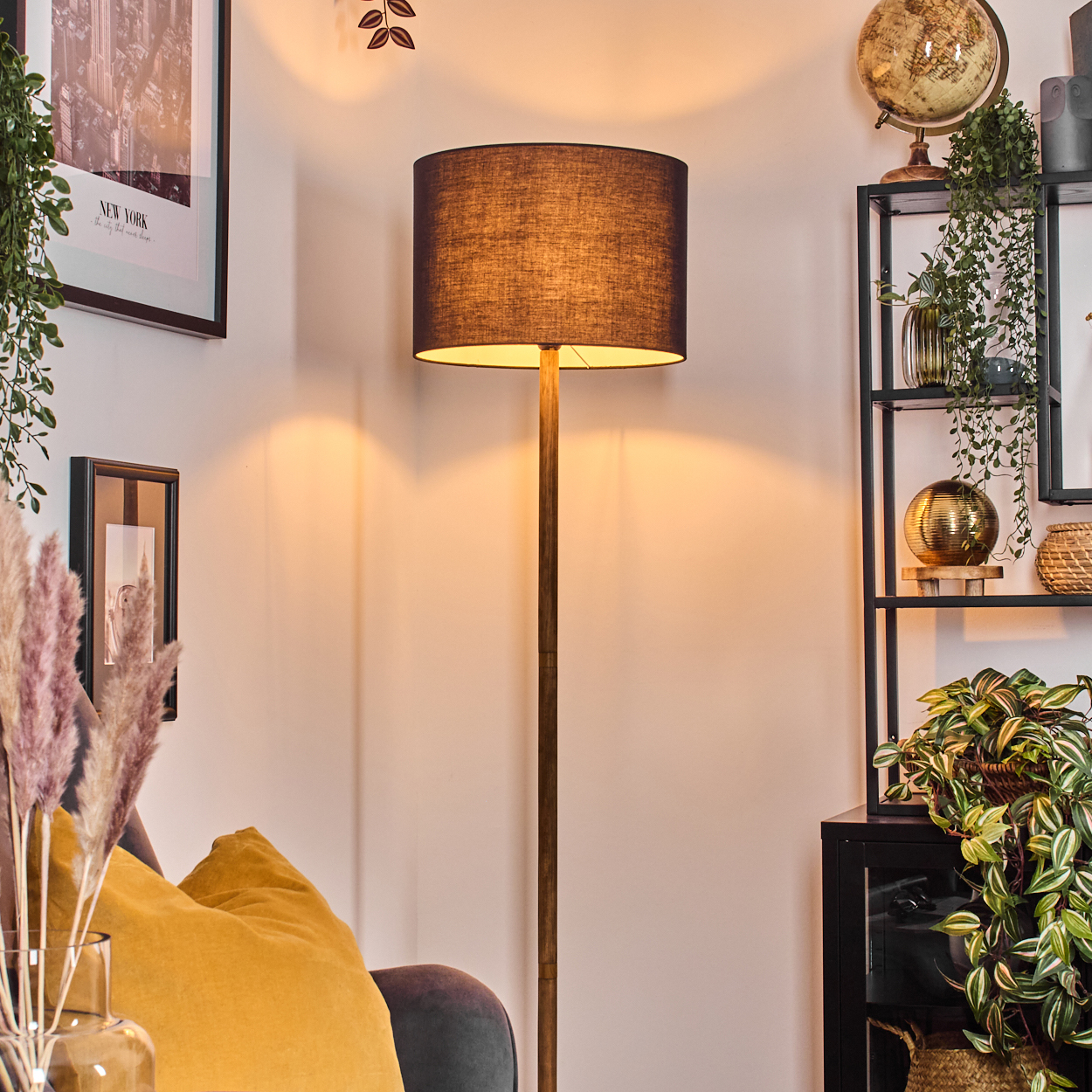 Eine Stehlampe beleuchtet einen Raum mit Pflanzen, Regalen, einem Globus und gerahmter Kunst. Ein gelbes Kissen liegt auf einem grauen Sofa.