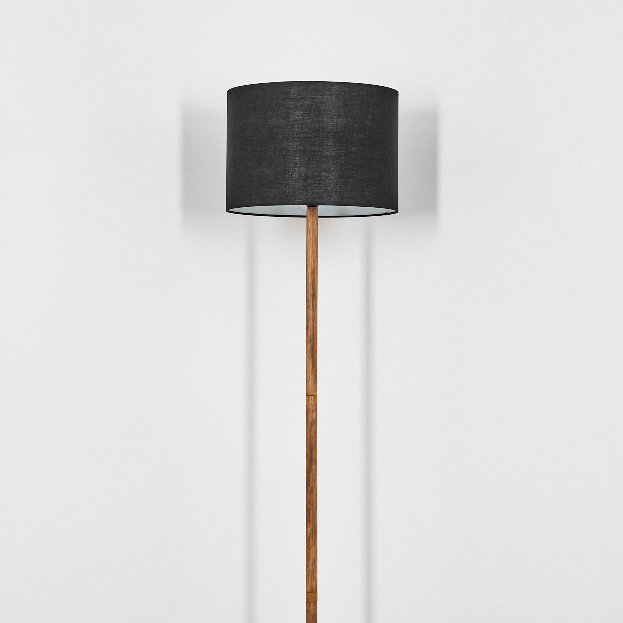 Eine hohe Lampe mit schwarzem Lampenschirm und Holzstange steht vor einer weißen Wand und wirft einen Schatten.