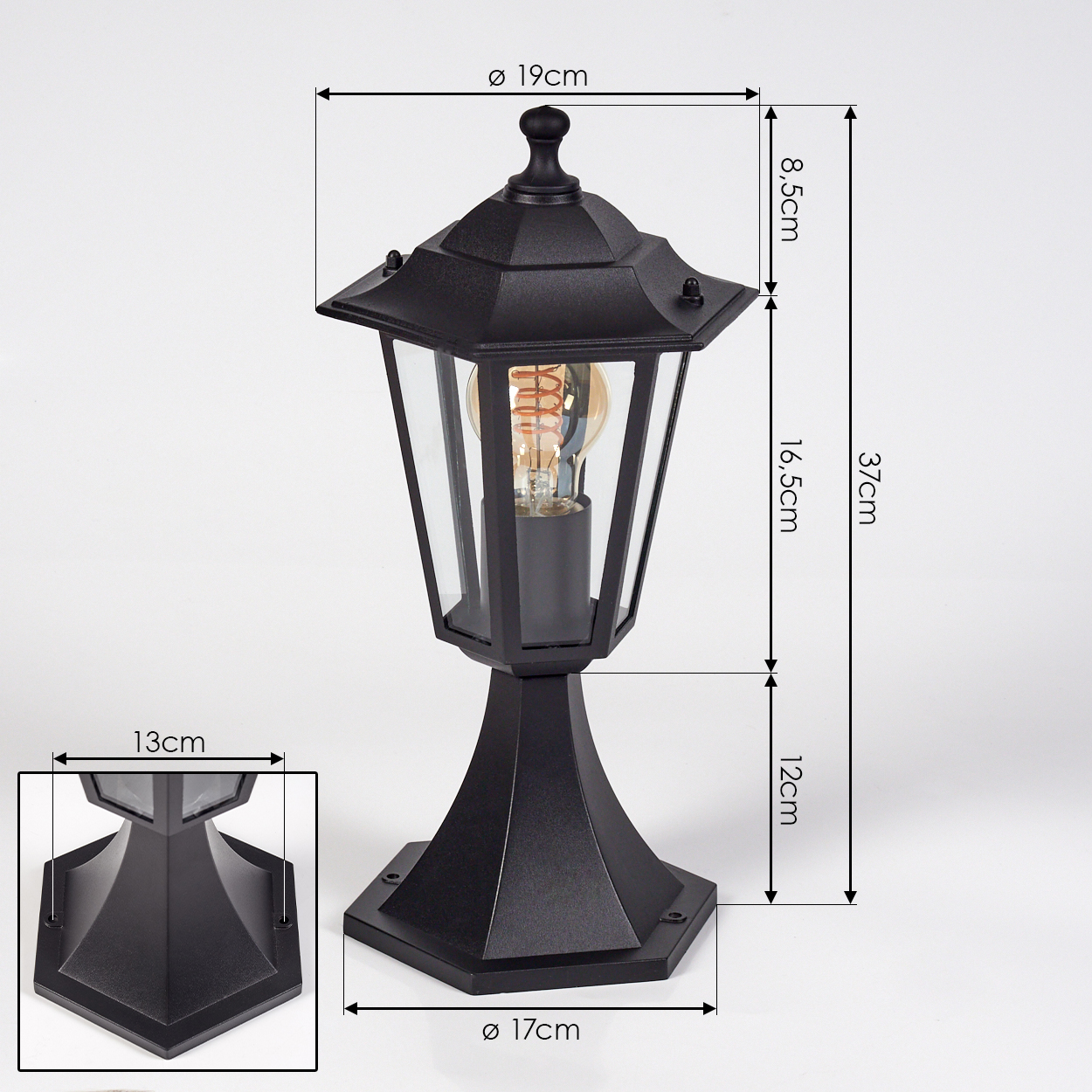 Eine schwarze Außenlampe mit beschrifteten Abmessungen. Sie hat ein Glasgehäuse, eine Glühbirne und eine dekorative Spitze. Der Sockel ist ebenfalls schwarz.