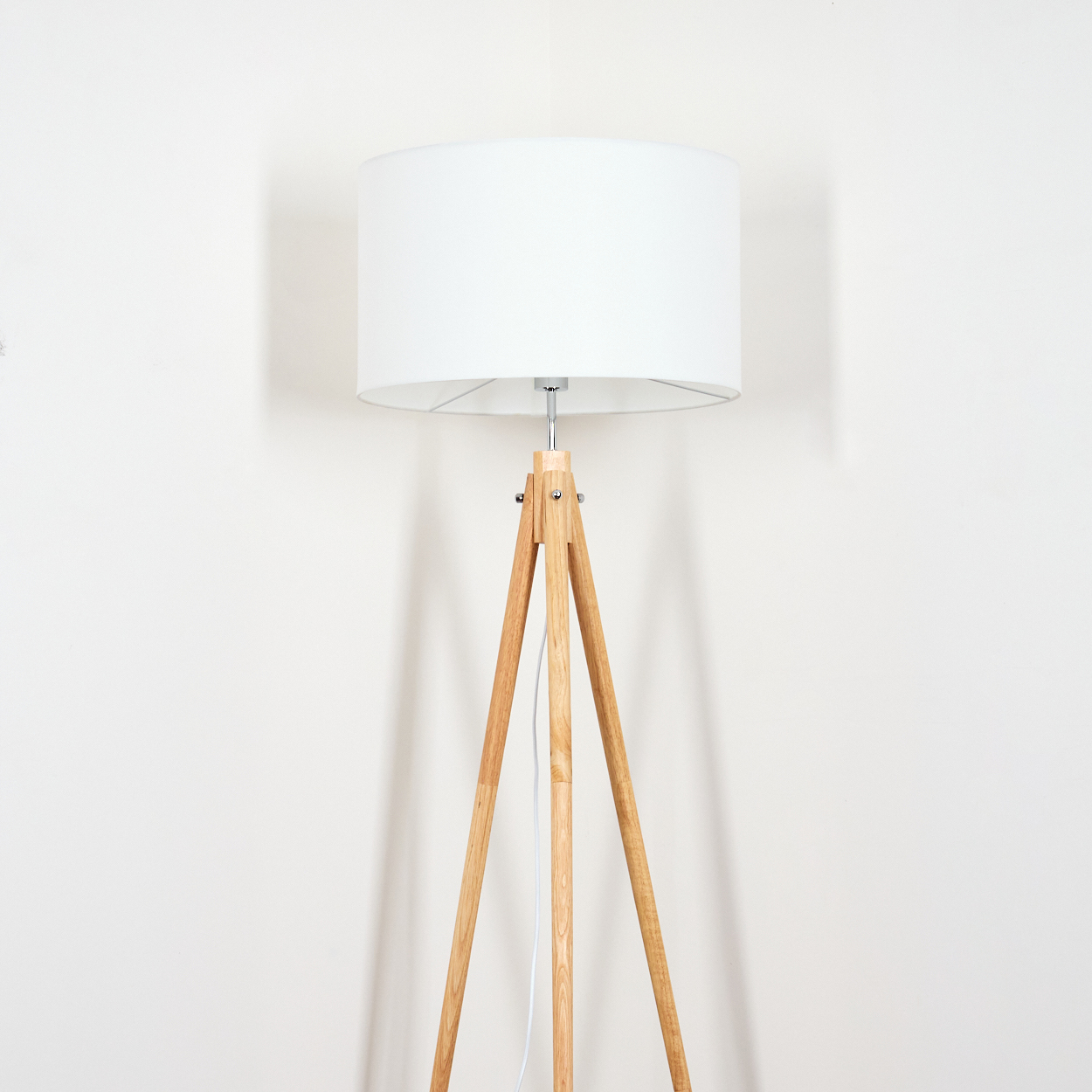 Eine Stehlampe mit hölzernem Stativ und weißem Lampenschirm steht vor einer weißen Wand. Die Beine der Lampe sind hellbraun.