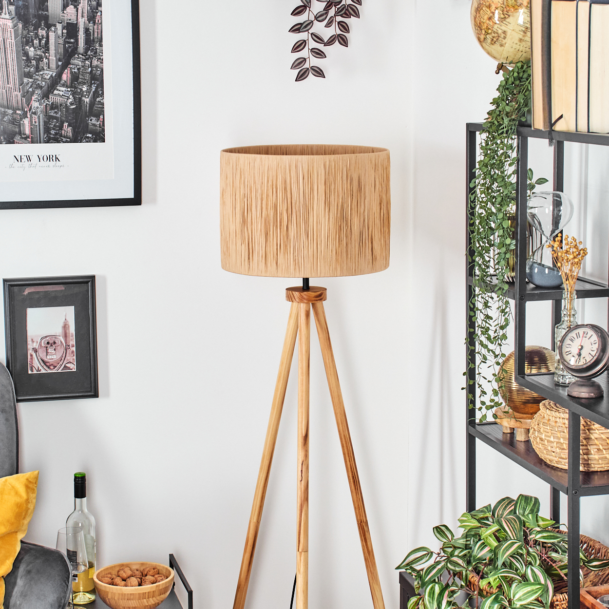Eine hölzerne Stativlampe mit beigem Lampenschirm steht in einem Raum. Regale mit Dekoration und ein New York-Poster sind im Hintergrund.
