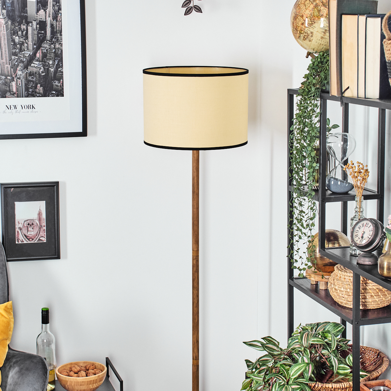 Eine Stehlampe mit beigem Schirm und schwarzem Rand steht vor einem Regal mit Dekoration. Eine New Yorker Skyline ist an der Wand.