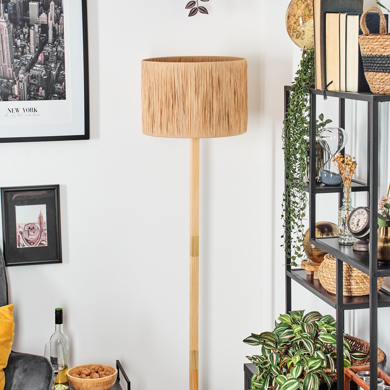 Eine Stehlampe mit beigem Schirm steht in einem Raum. Rechts ein Regal mit Büchern und Pflanzen. Bilder hängen an der Wand.