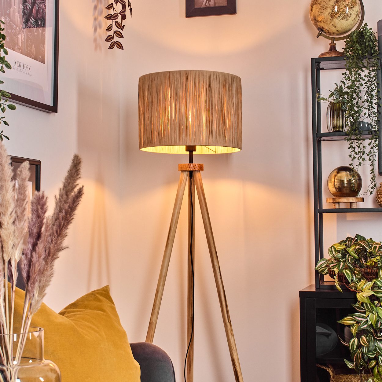 Eine hölzerne Stativlampe mit beigem Schirm beleuchtet ein Wohnzimmer mit Regal, Pflanzen und einem gelben Kissen.