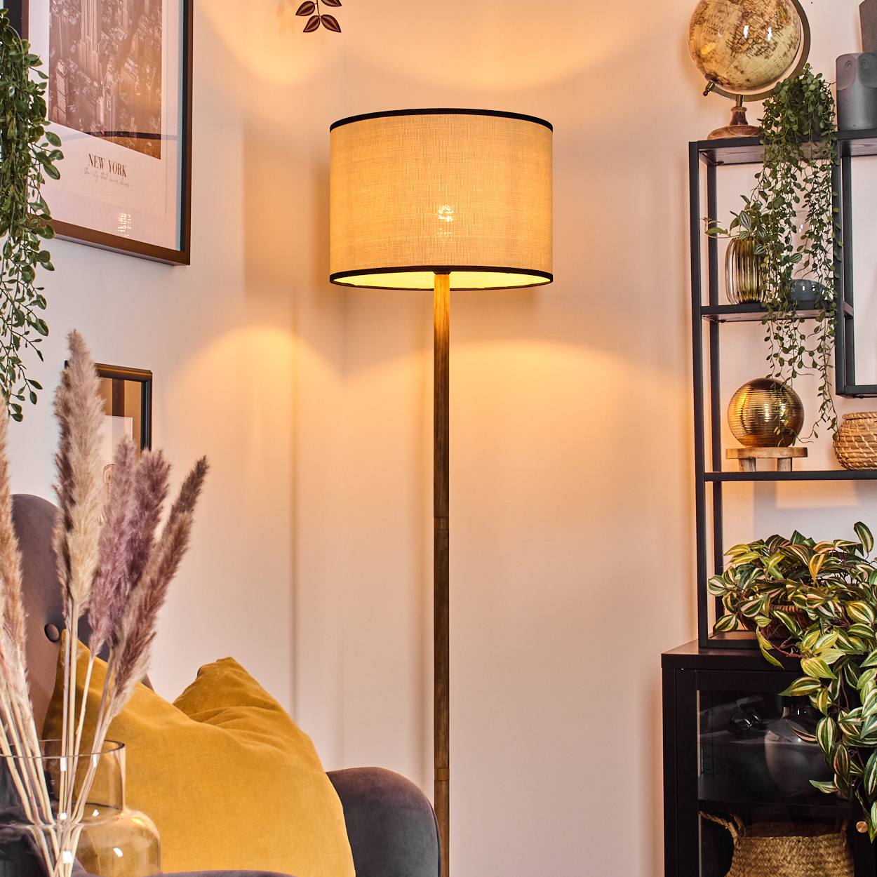 Eine Stehlampe mit beigem Schirm steht neben einem Regal mit Dekorationen. Ein gelbes Kissen und Pampasgras sind im Vordergrund.