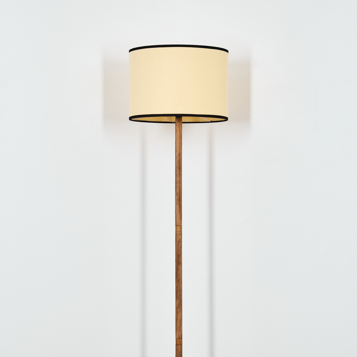 Eine hölzerne Stehlampe mit cremefarbenem Lampenschirm und schwarzem Rand steht vor einer weißen Wand. Ein Schatten wird geworfen.