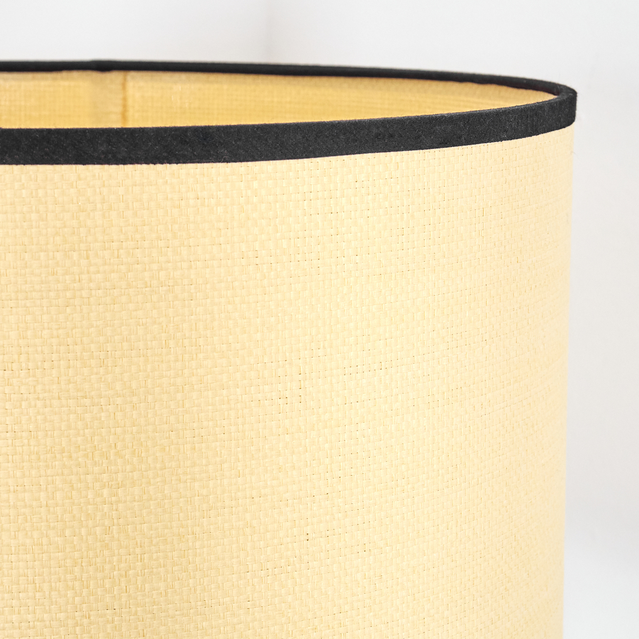 Nahaufnahme eines Lampenschirms. Er hat eine gewebte beige Außenseite mit einem schwarzen Band am oberen Rand. Der Hintergrund ist weiß.