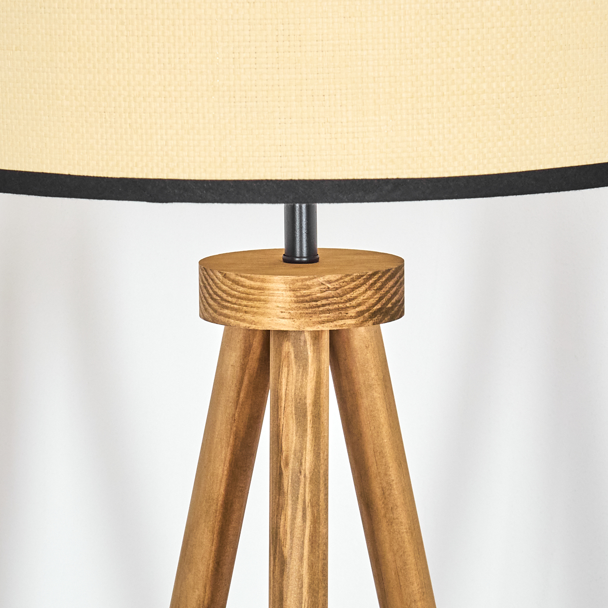 Nahaufnahme einer Holzstativlampe. Sie hat einen beigefarbenen Lampenschirm mit schwarzem Rand. Die Beine sind aus Holz, der Hintergrund ist weiß.