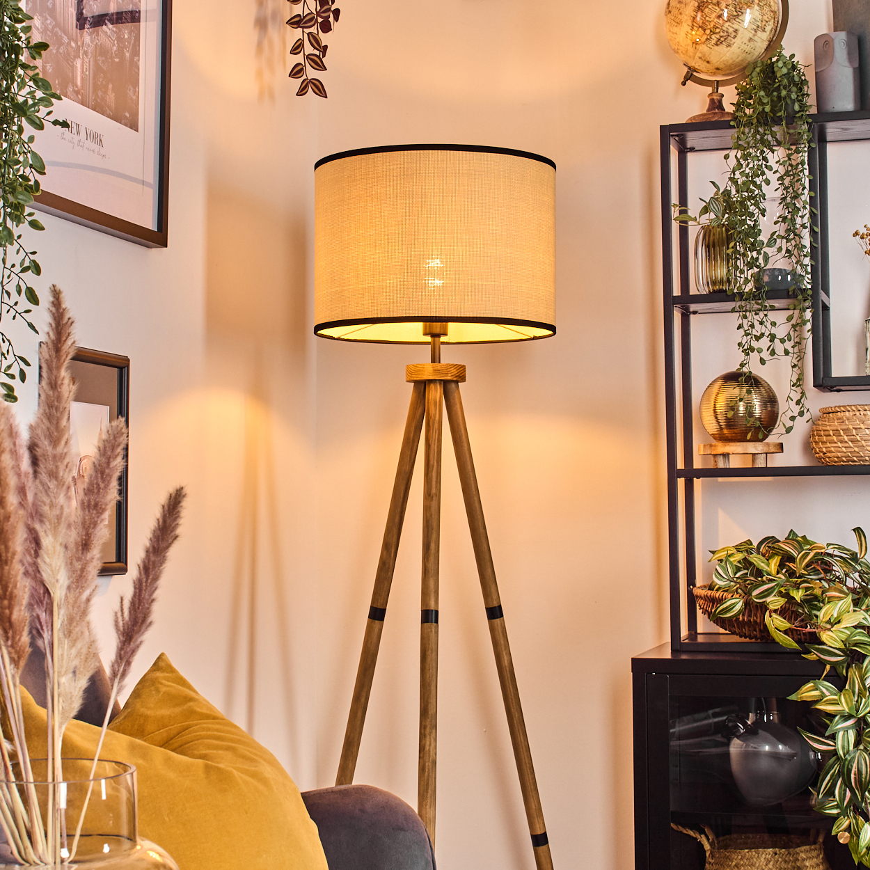 Eine beleuchtete Stehlampe mit Stativ steht in einer Zimmerecke. Dahinter ein schwarzes Regal mit Pflanzen und Dekoration. Im Vordergrund ein Sofa mit gelben Kissen.