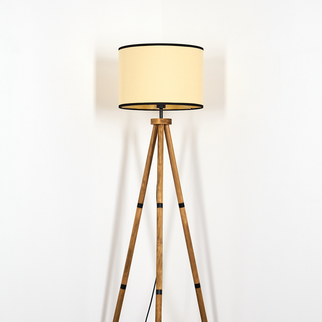 Eine hölzerne Stativ-Stehlampe mit beigem Lampenschirm vor einer weißen Wand. Schwarze Akzente an den Holzbeinen und dem Schirm.