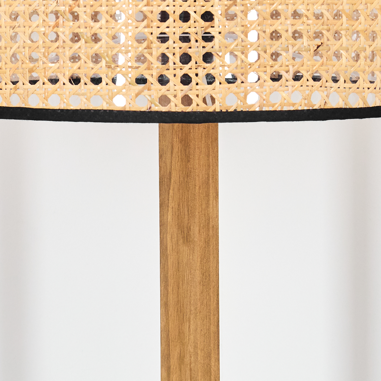 Nahaufnahme einer Lampe. Gewebter Rattanschatten mit schwarzem Rand sitzt über einem Holzpfosten. Weiße Wand im Hintergrund.